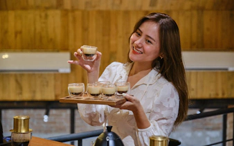Woman holding tray of Vietnamese egg coffee glasses in a cafe setting.