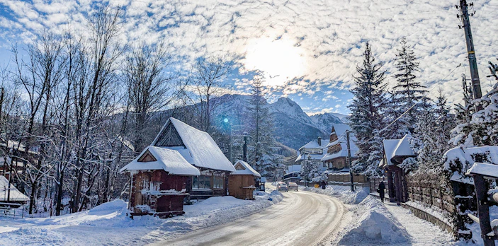 Wooden houses covered in snow along a road in Zakopane, with mountains in the background.