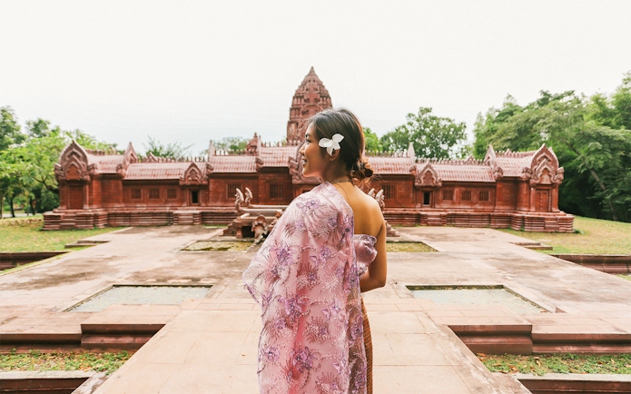 Woman in traditional attire at Ancient City, Thailand, with red temple in background.