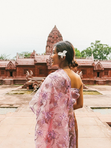 Woman in traditional attire at Ancient City, Thailand, with red temple in background.
