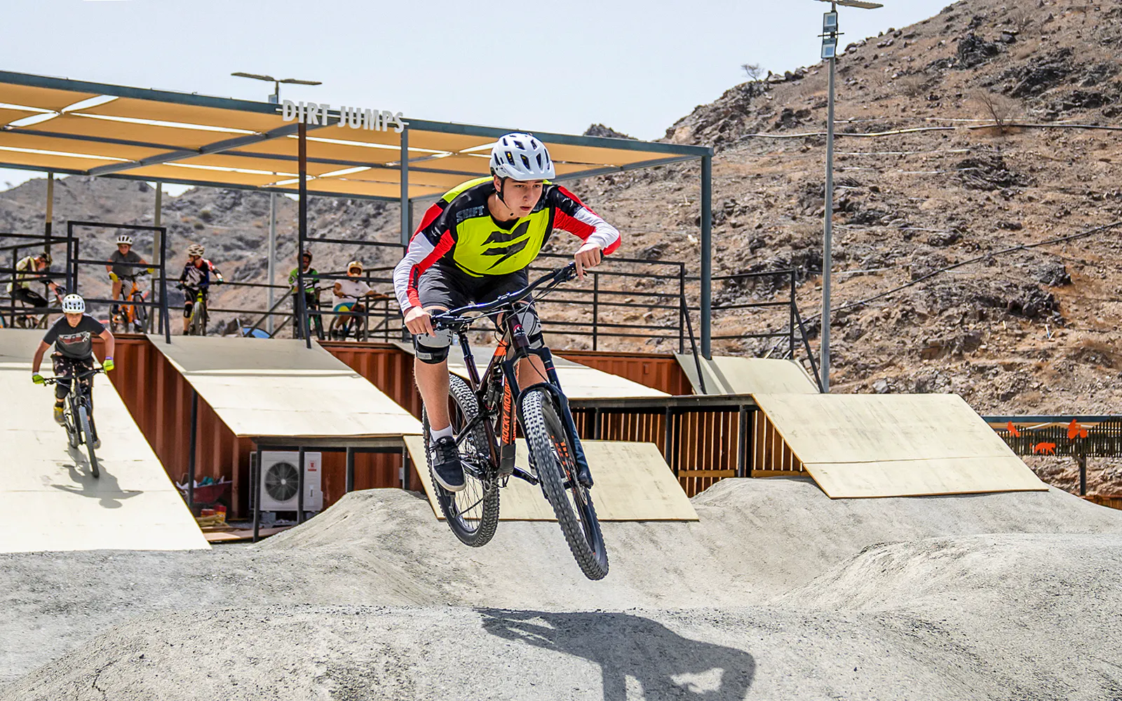 A mountain biker clears a dirt jump at Fujairah Adventure Park with rocky hills behind.