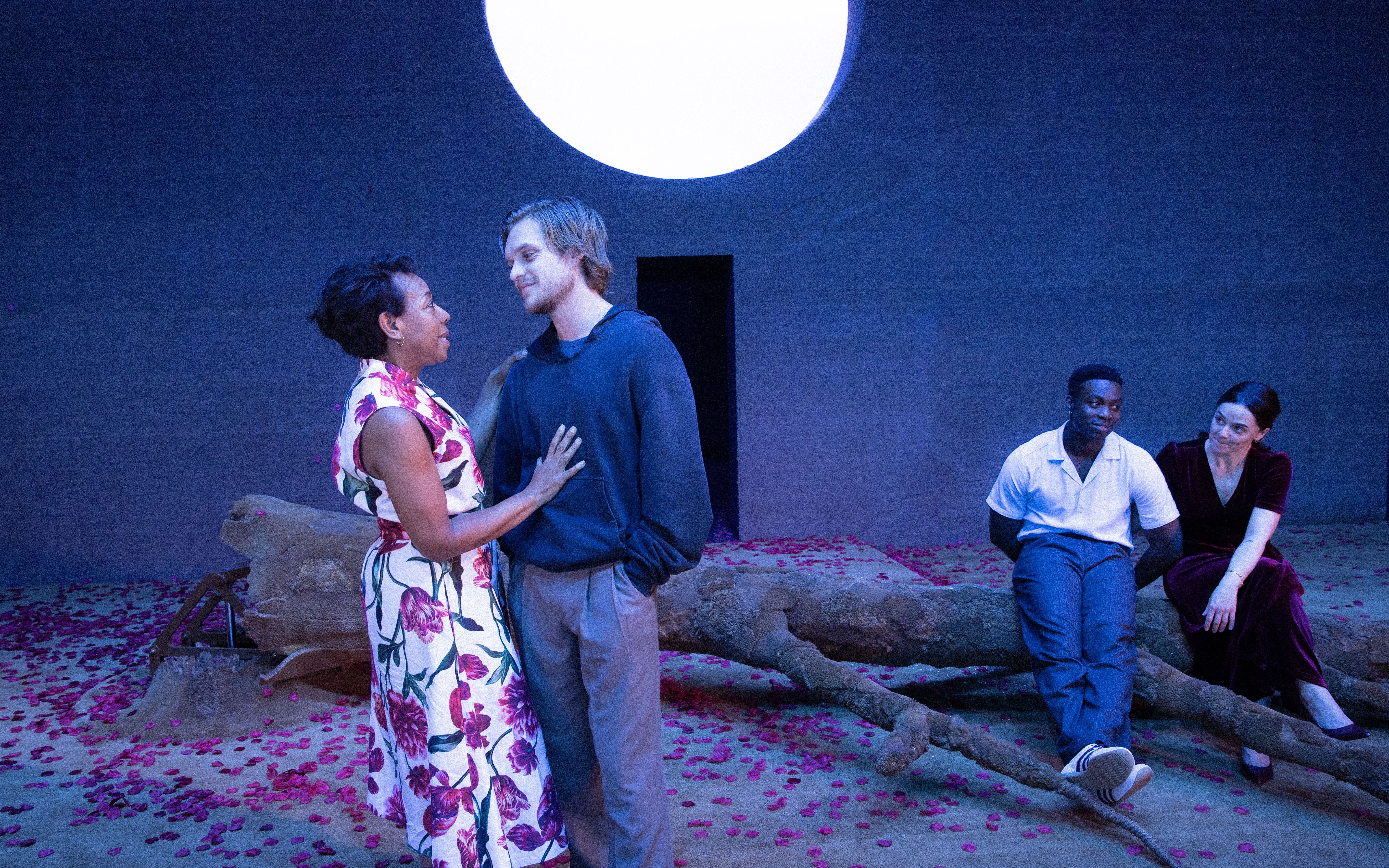 Stage scene from All My Sons West End show with actors interacting on a set with a large moon backdrop.