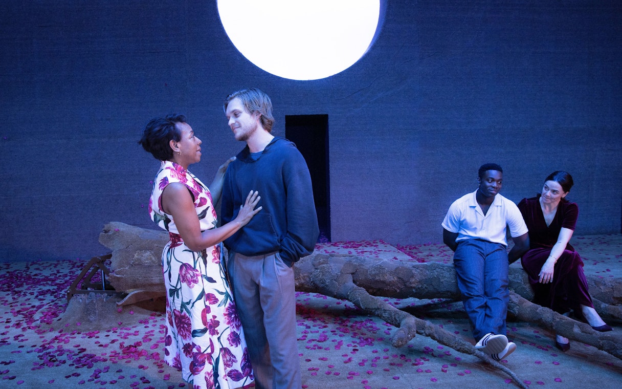 Stage scene from All My Sons West End show with actors interacting on a set with a large moon backdrop.