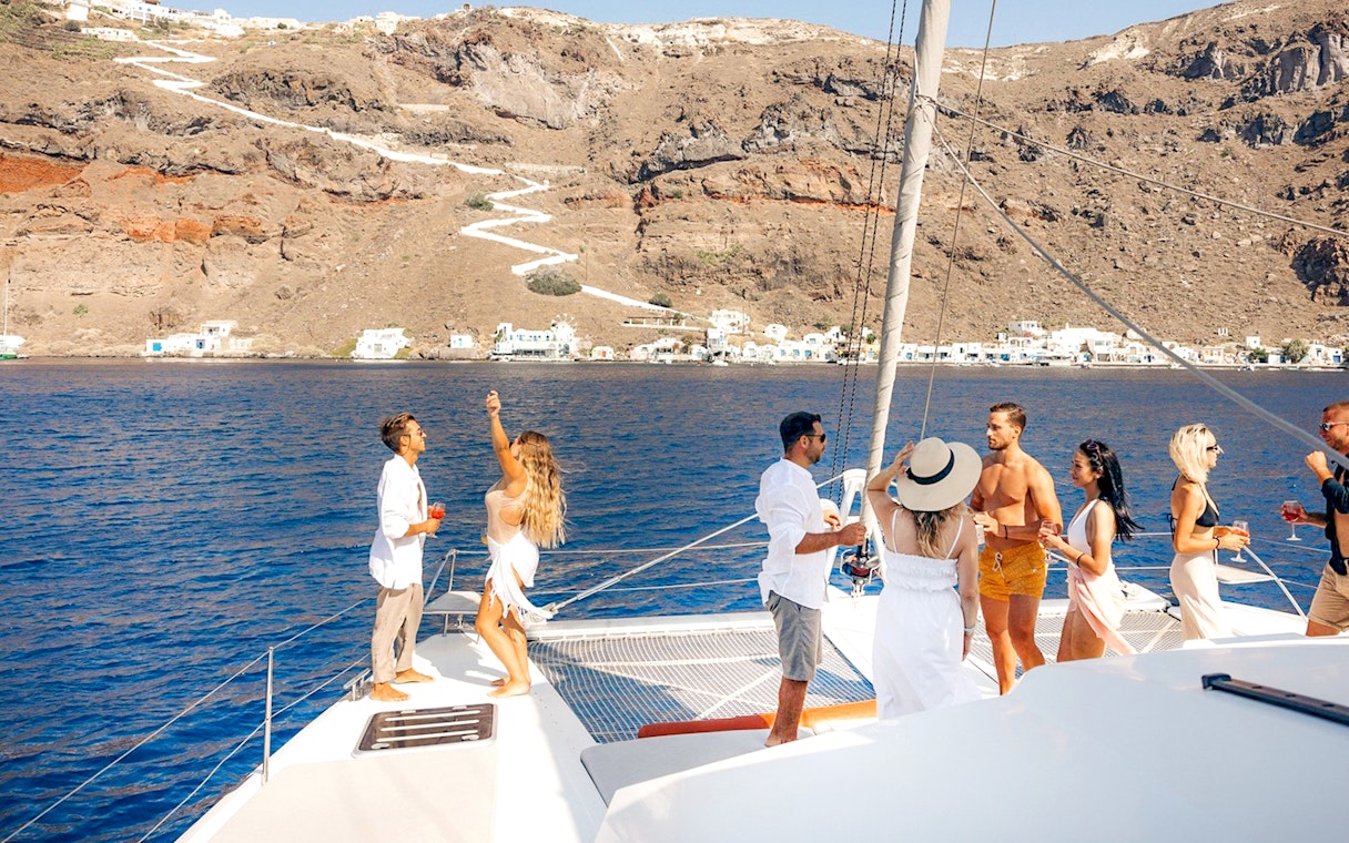 Guests enjoying drinks on a catamaran cruise in Santorini with scenic coastline views.