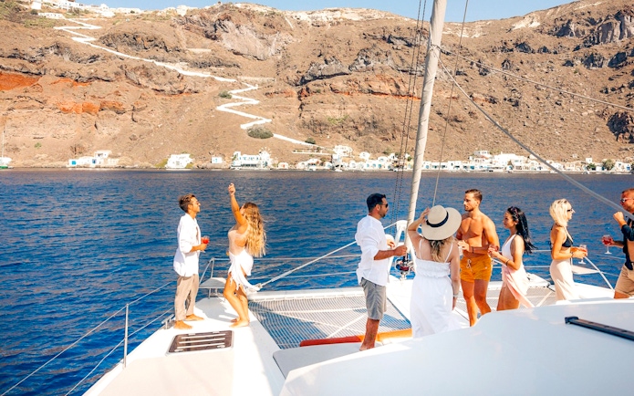 Guests enjoying drinks on a catamaran cruise in Santorini with scenic coastline views.