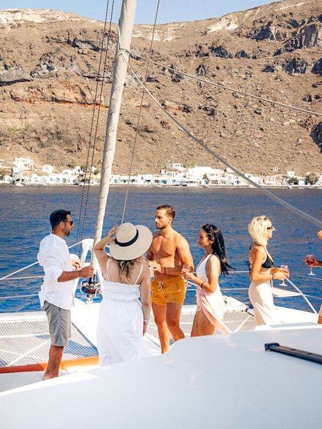 Guests enjoying drinks on a catamaran cruise in Santorini with scenic coastline views.
