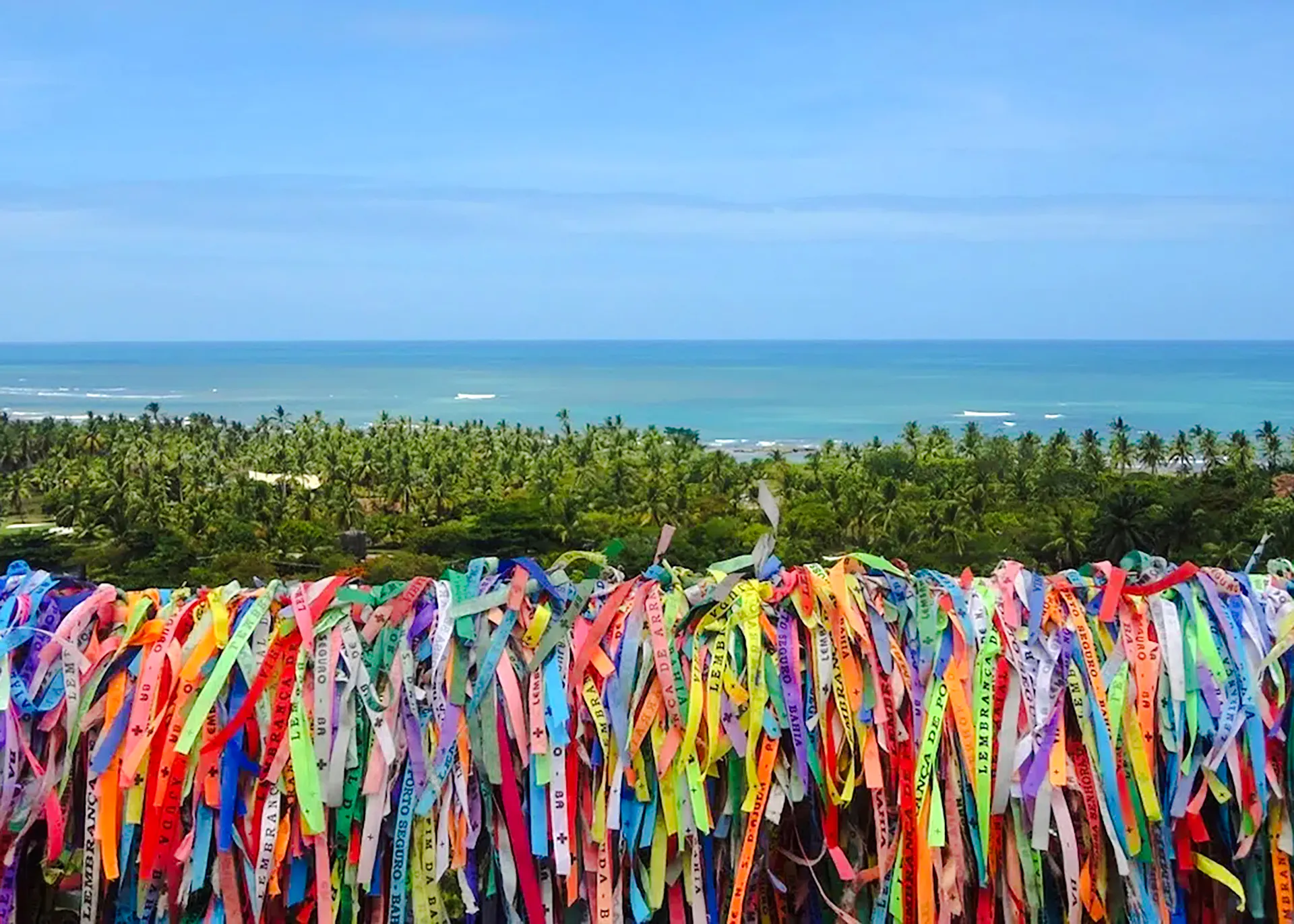 Colorful ribbons tied on a fence overlooking palm trees and ocean in Arraial d'Ajuda, Porto Seguro.