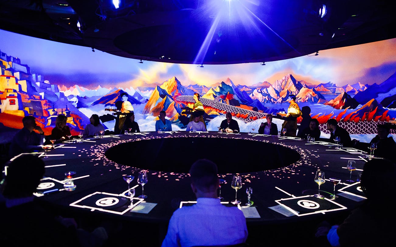 Guests seated around a circular table at IMAGINARY ART show, KRASOTA Dubai, with vibrant mountain mural.