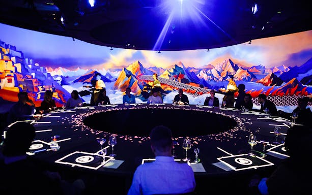 Guests seated around a circular table at IMAGINARY ART show, KRASOTA Dubai, with vibrant mountain mural.