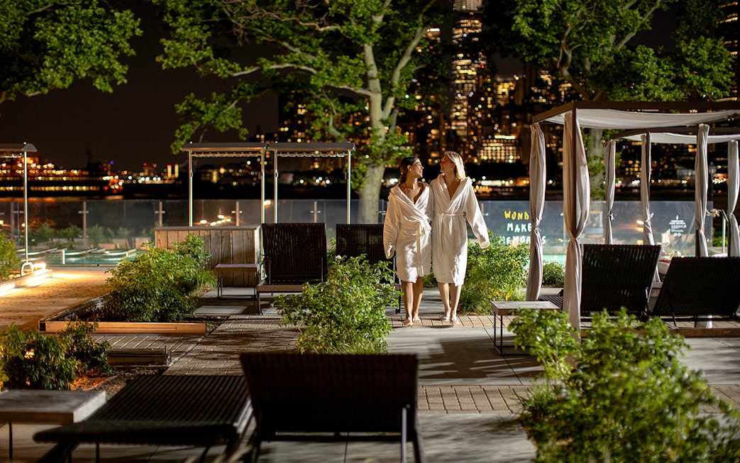 Couple in robes walking at night in QC New York spa garden with city skyline.