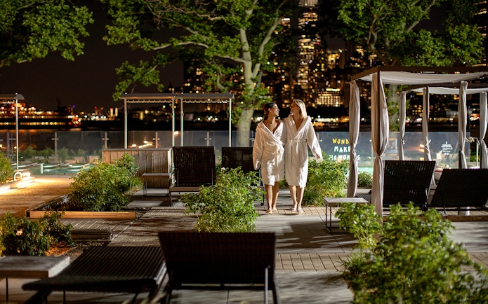 Couple in robes walking at night in QC New York spa garden with city skyline.