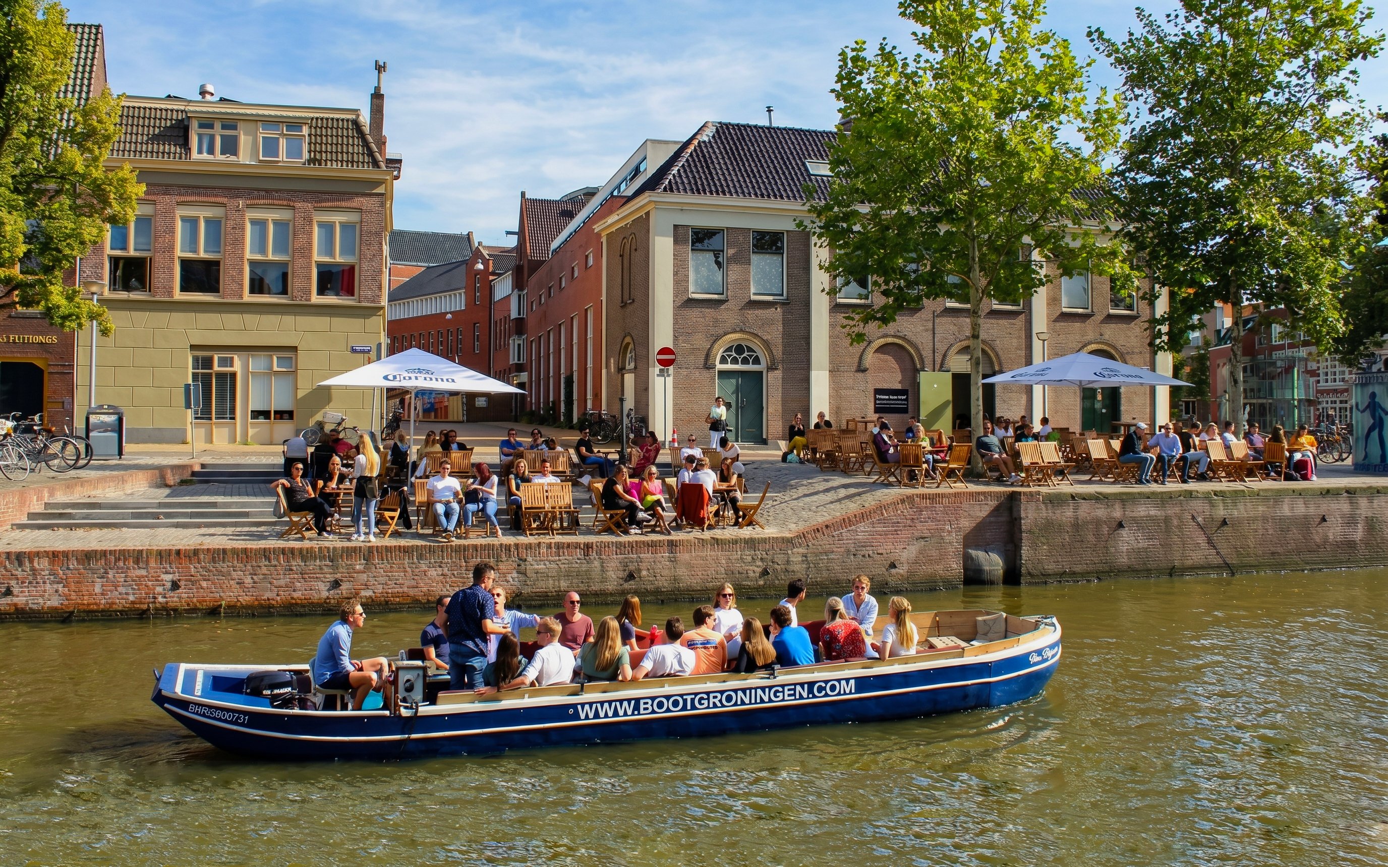 Boat tour on Groningen canal with people enjoying outdoor cafes along the waterfront.