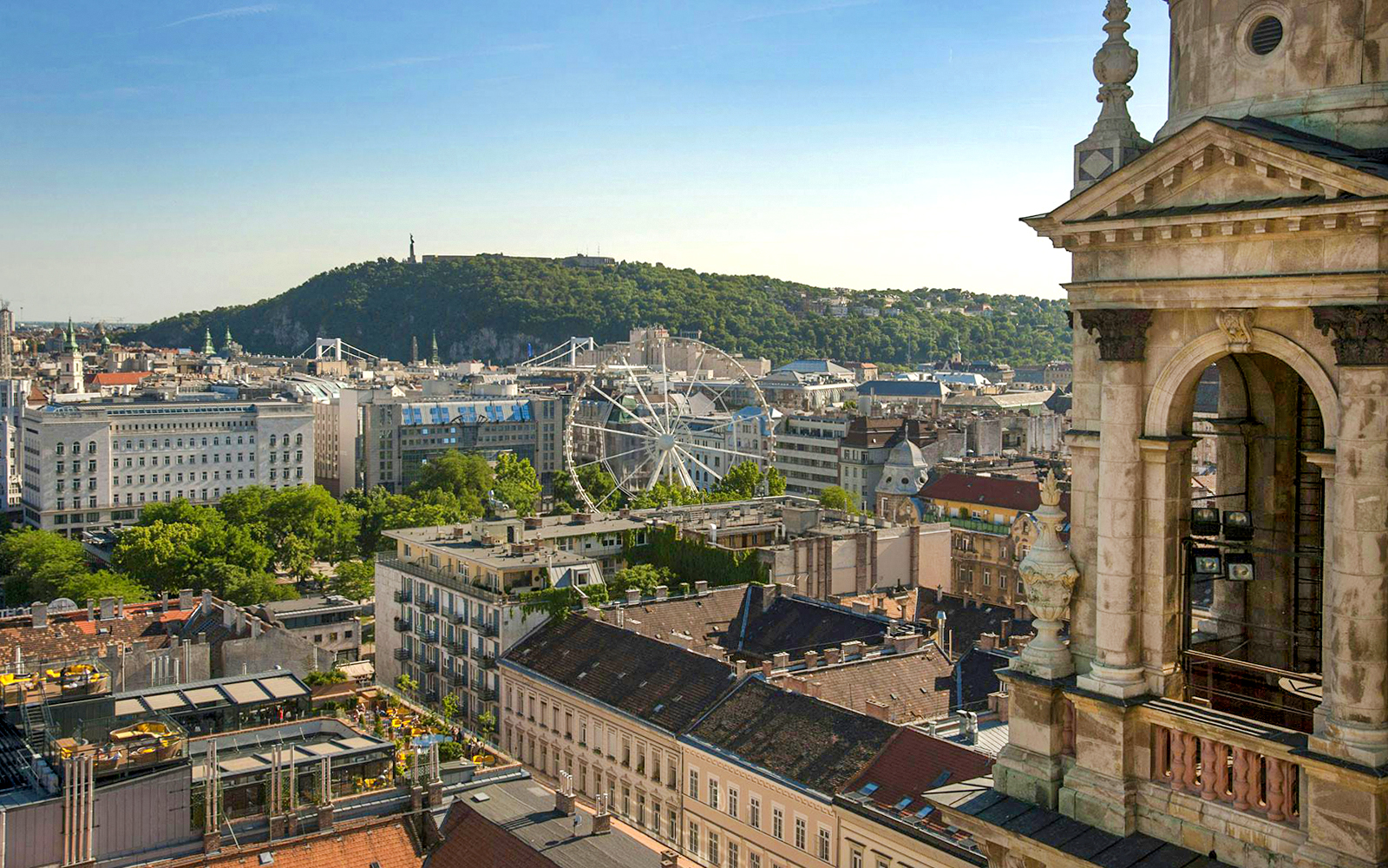 View of Budapest cityscape from St. Stephen's Basilica, featuring a Ferris wheel and Gellért Hill.