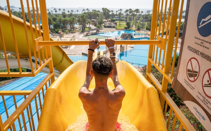 Person on a yellow water slide at Aquopolis Costa Daurada, Tarragona, with pools and palm trees in view.