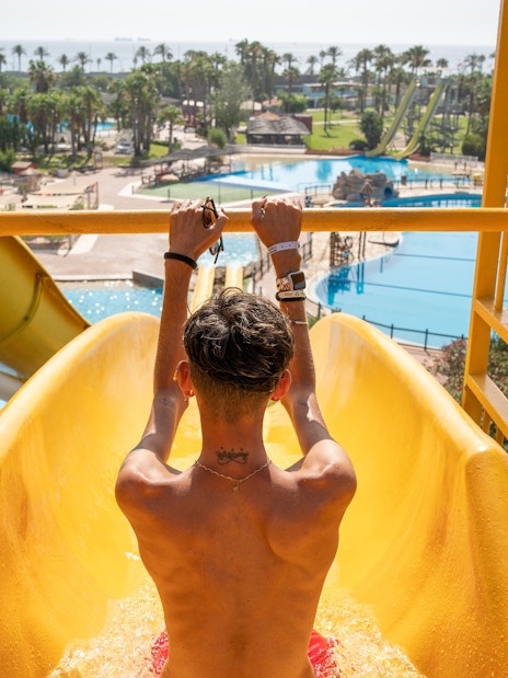 Person on a yellow water slide at Aquopolis Costa Daurada, Tarragona, with pools and palm trees in view.