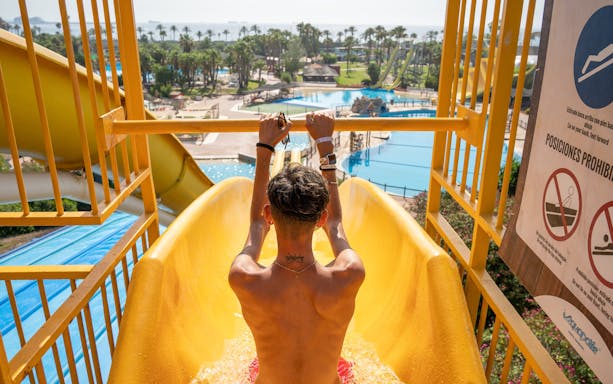 Person on a yellow water slide at Aquopolis Costa Daurada, Tarragona, with pools and palm trees in view.