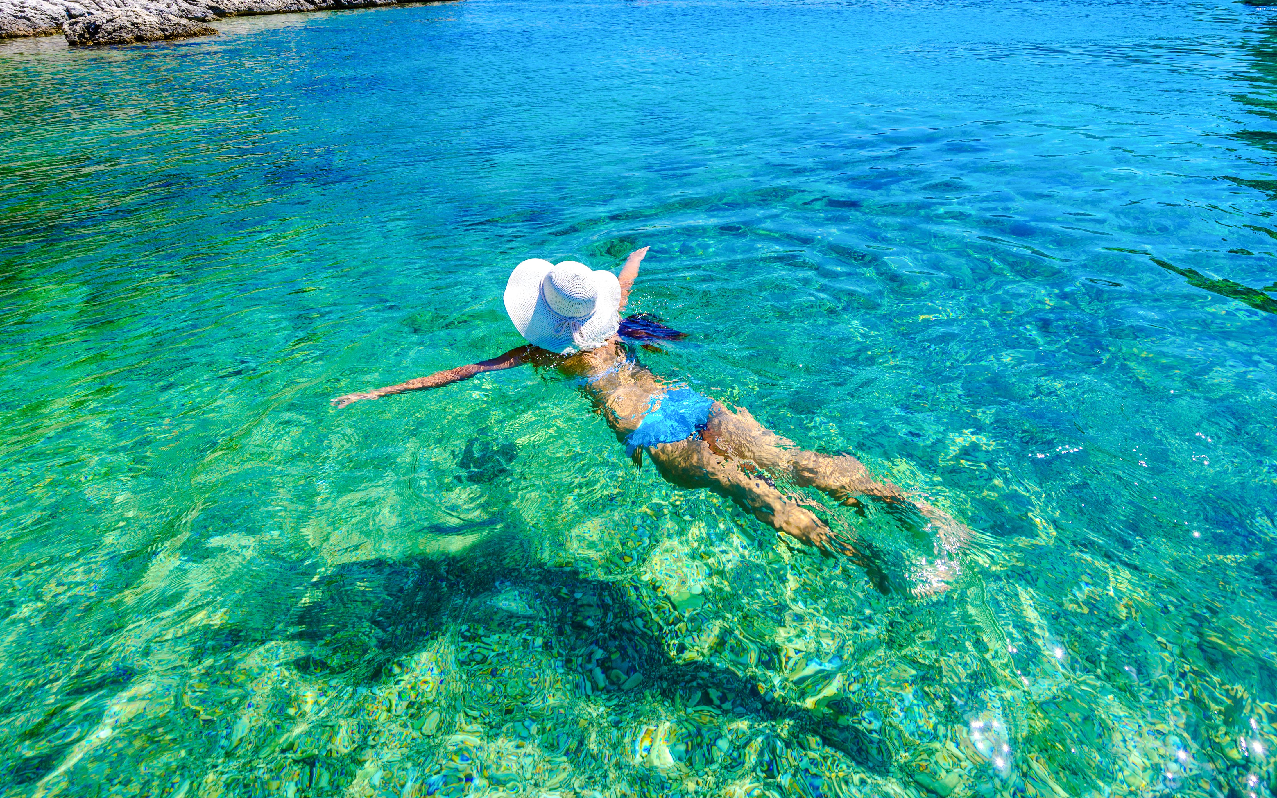 Woman swimming in clear turquoise bay wearing a sun hat.