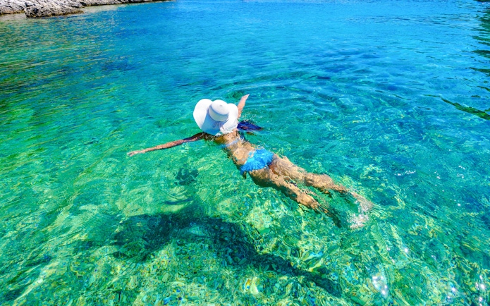 Woman swimming in clear turquoise bay wearing a sun hat.