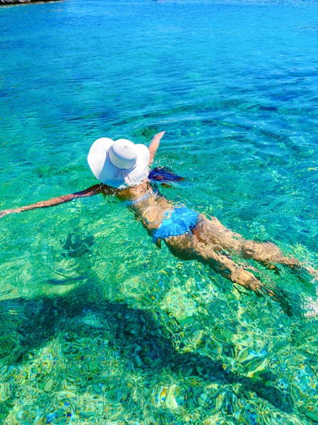 Woman swimming in clear turquoise bay wearing a sun hat.