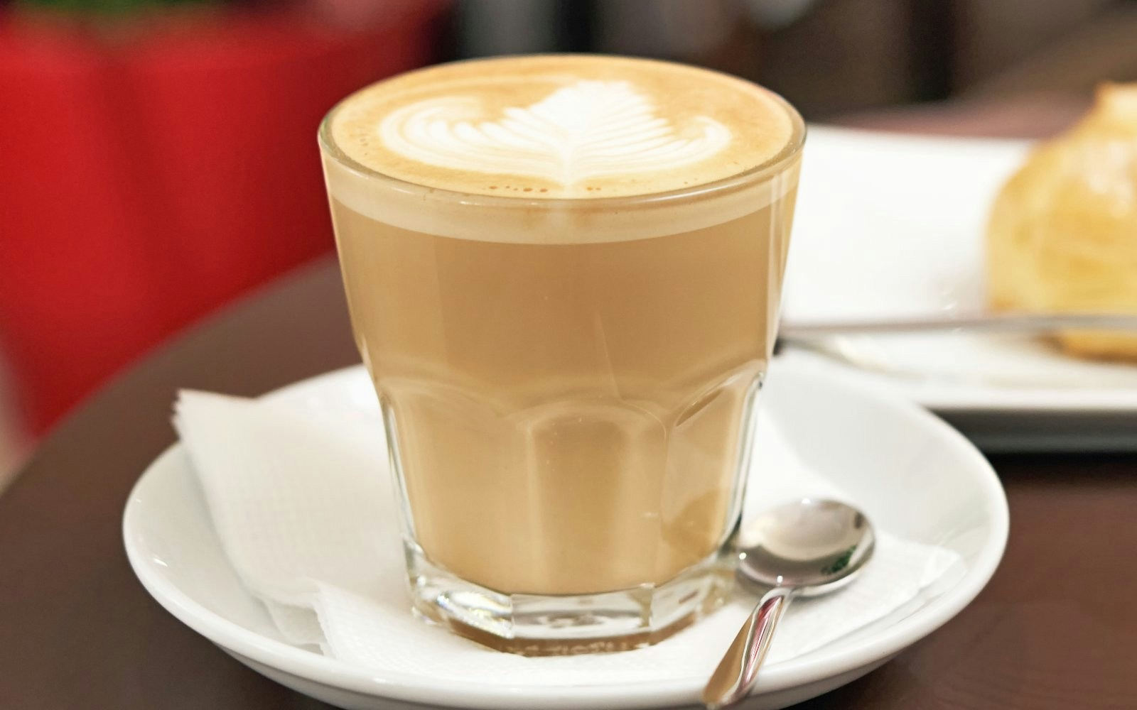 Flat white coffee in a glass with white foam, eclair cake in the background.