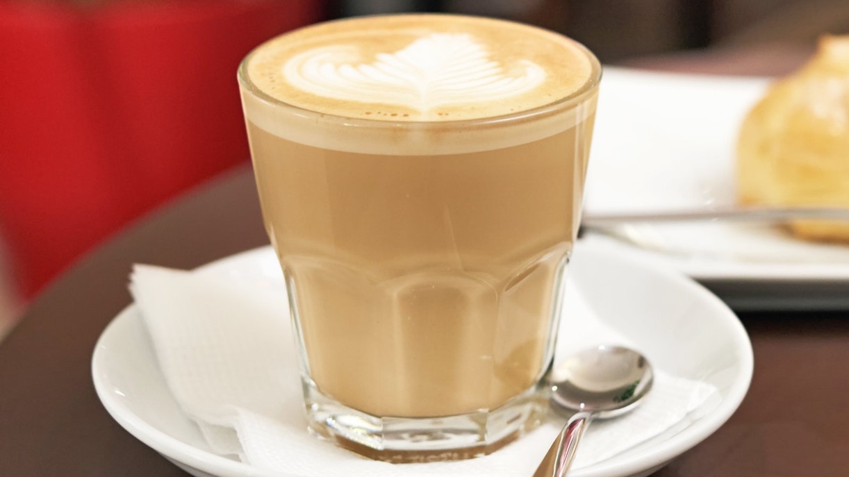 Flat white coffee in a glass with white foam, eclair cake in the background.