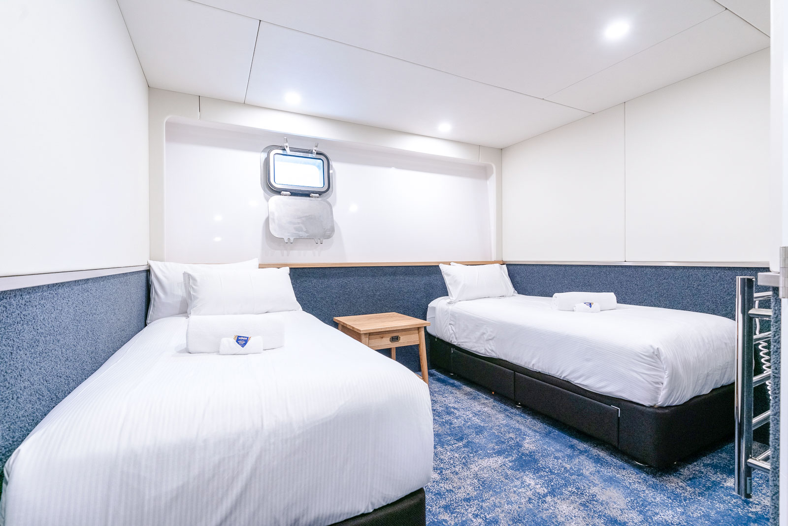 Twin cabin interior with two beds, a mirror, and a heated towel rack on a ship.