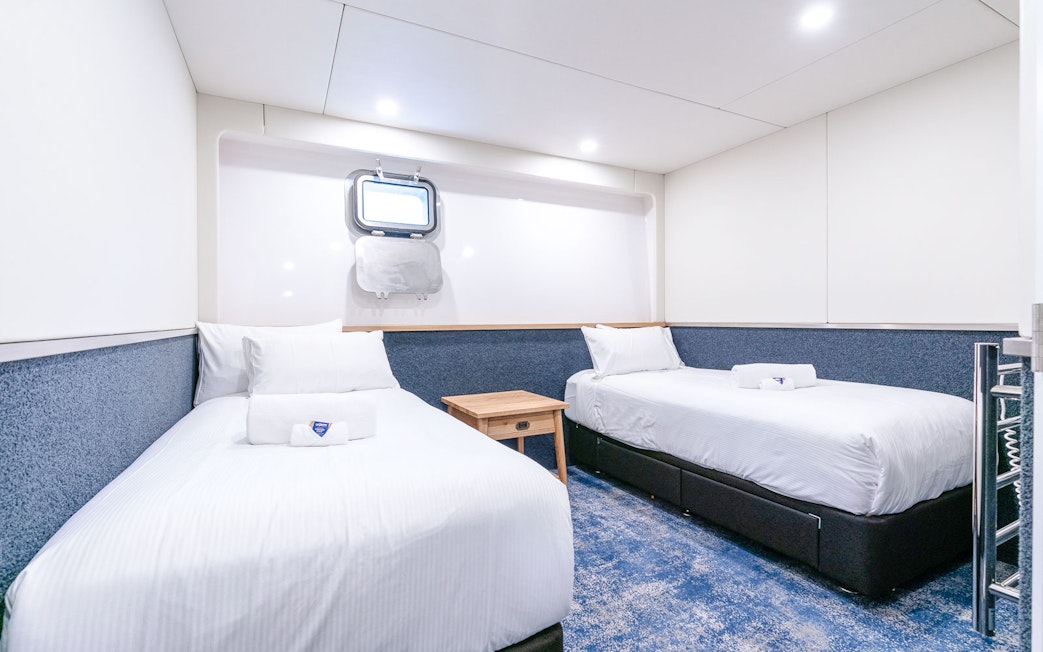 Twin cabin interior with two beds, a mirror, and a heated towel rack on a ship.