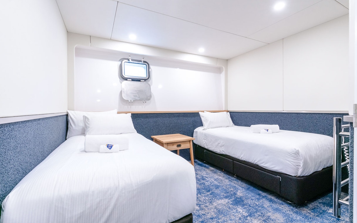 Twin cabin interior with two beds, a mirror, and a heated towel rack on a ship.