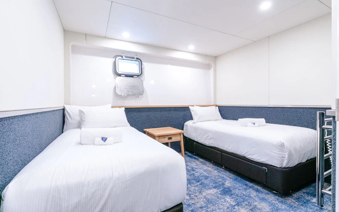Twin cabin interior with two beds, a mirror, and a heated towel rack on a ship.