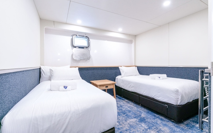 Twin cabin interior with two beds, a mirror, and a heated towel rack on a ship.