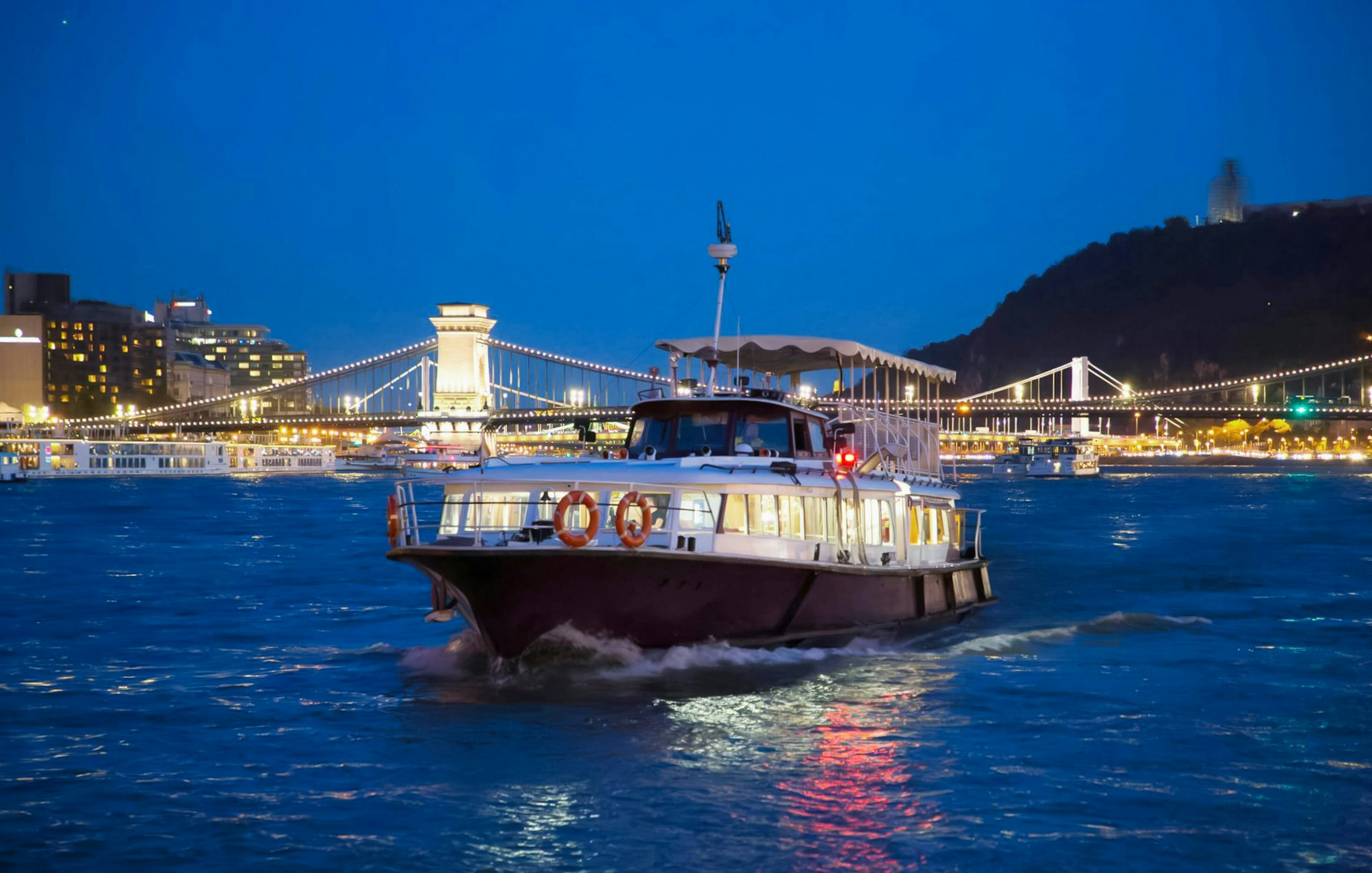 Cruise boat on the Danube River with Budapest Chain Bridge illuminated at night.