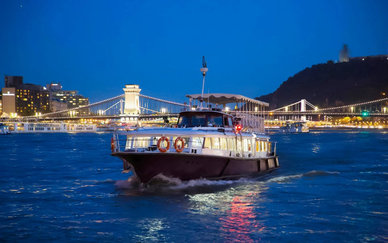 Cruise boat on the Danube River with Budapest Chain Bridge illuminated at night.