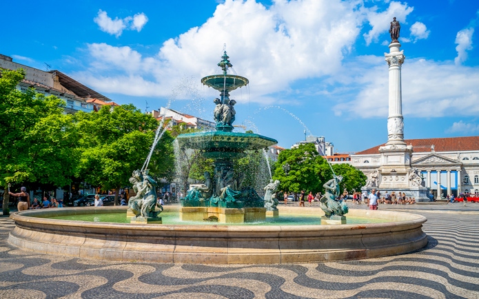 Fountain and column in Rossio Square, Lisbon, with people and trees in the background.