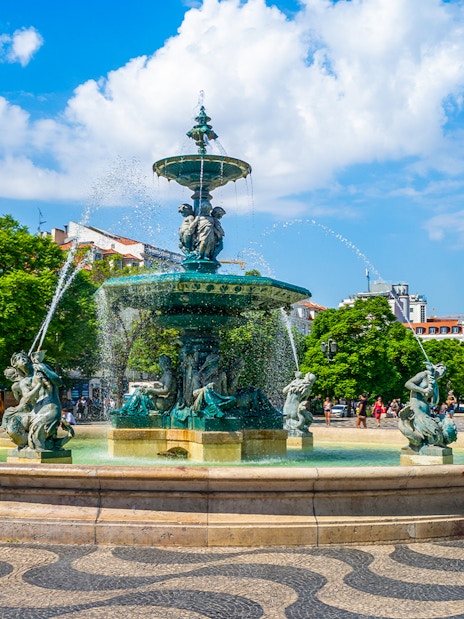 Fountain and column in Rossio Square, Lisbon, with people and trees in the background.