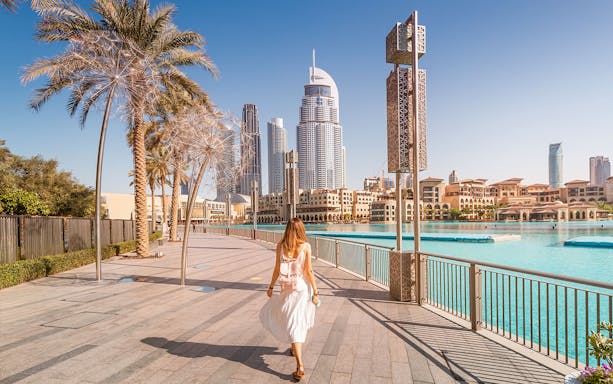 Woman walking along Dubai waterfront with Burj Khalifa and city skyline in view.
