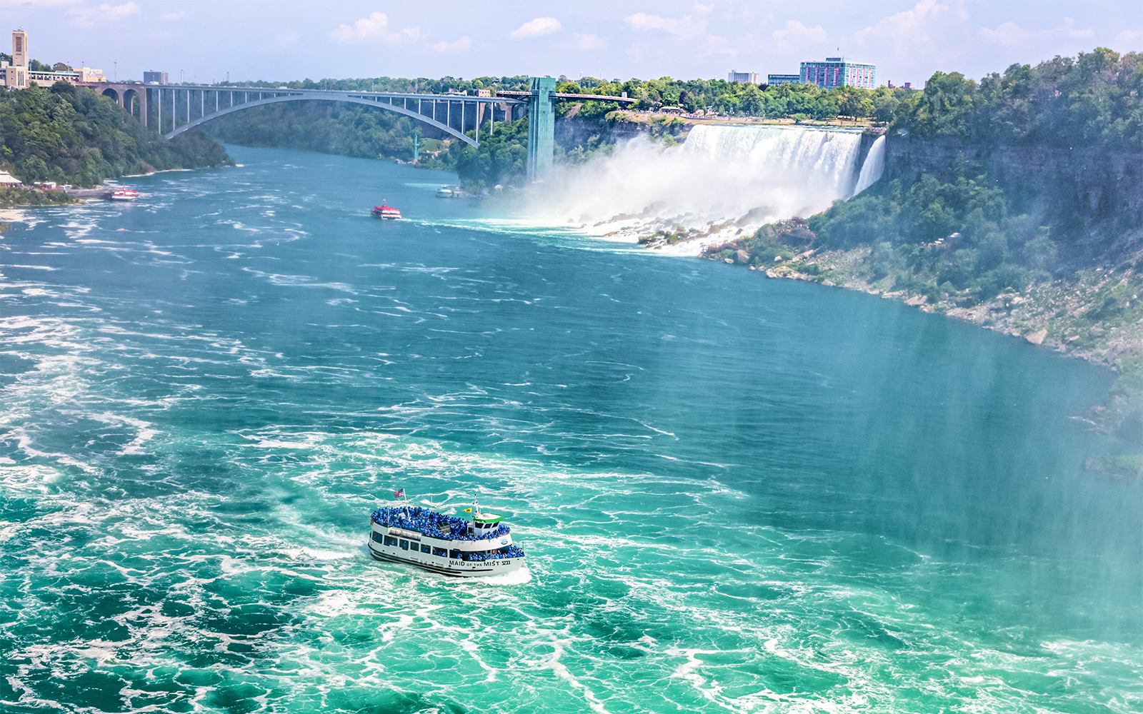 Maid of the Mist boat approaching Niagara Falls, USA with mist and bridge in background.