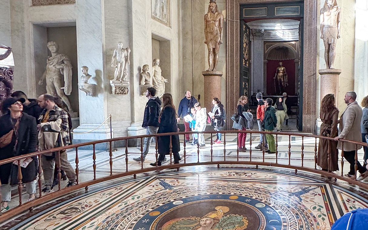 Visitors exploring the Round Hall in the Vatican Museums, Rome, with ancient statues and mosaic floor.