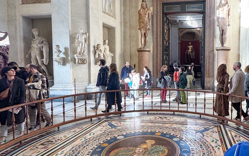 Visitors exploring the Round Hall in the Vatican Museums, Rome, with ancient statues and mosaic floor.