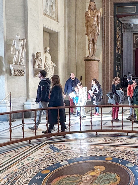Visitors exploring the Round Hall in the Vatican Museums, Rome, with ancient statues and mosaic floor.