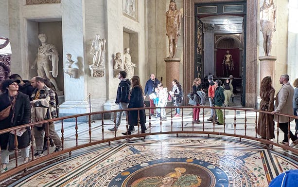 Visitors exploring the Round Hall in the Vatican Museums, Rome, with ancient statues and mosaic floor.