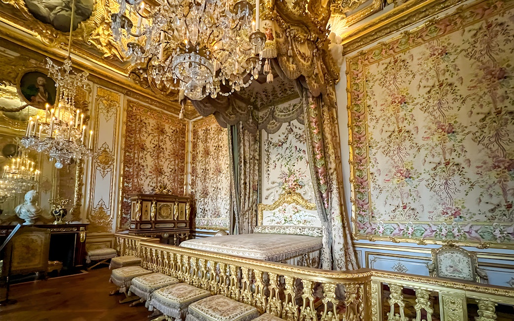 Queens apartment interior at Versailles Palace, France, with ornate bed, floral tapestries, and chandelier.
