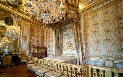 Queens apartment interior at Versailles Palace, France, with ornate bed, floral tapestries, and chandelier.