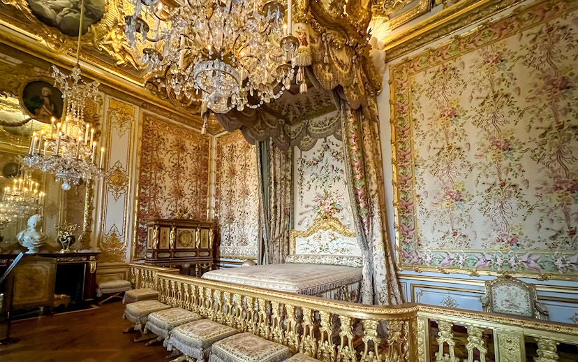Queens apartment interior at Versailles Palace, France, with ornate bed, floral tapestries, and chandelier.