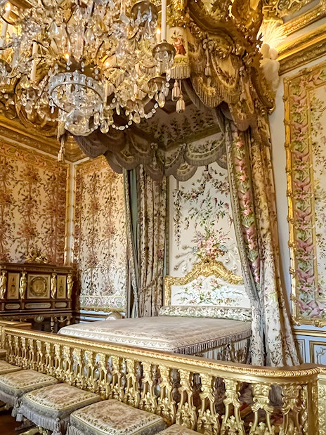 Queens apartment interior at Versailles Palace, France, with ornate bed, floral tapestries, and chandelier.