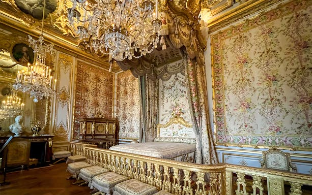 Queens apartment interior at Versailles Palace, France, with ornate bed, floral tapestries, and chandelier.