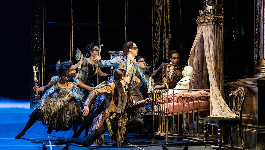 Performers in elaborate costumes surround a crib in Matthew Bourne's Sleeping Beauty production.