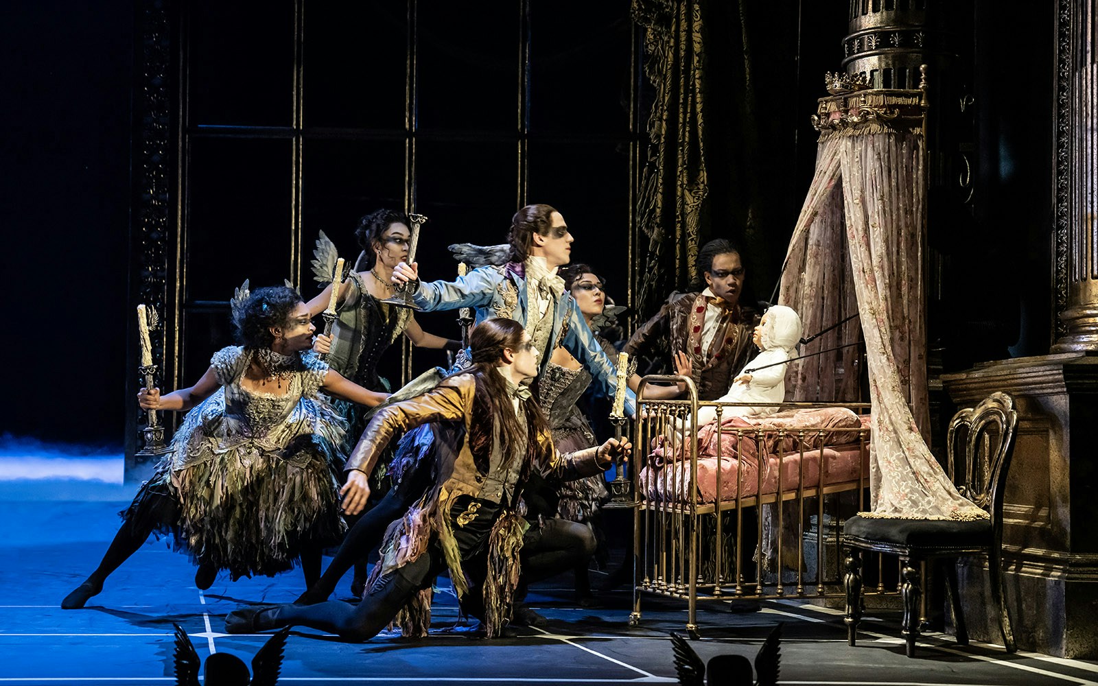Performers in elaborate costumes surround a crib in Matthew Bourne's Sleeping Beauty production.