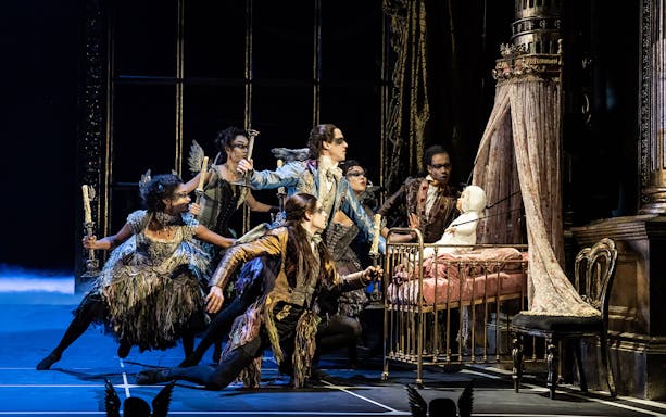 Performers in elaborate costumes surround a crib in Matthew Bourne's Sleeping Beauty production.