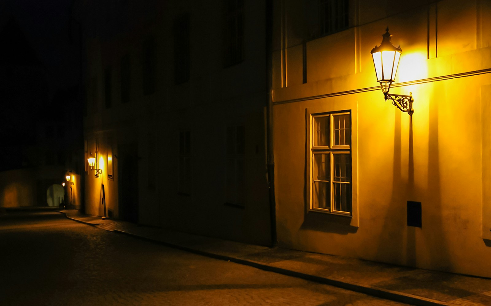 Old lanterns lighting a narrow alleyway in a historic European city at night.