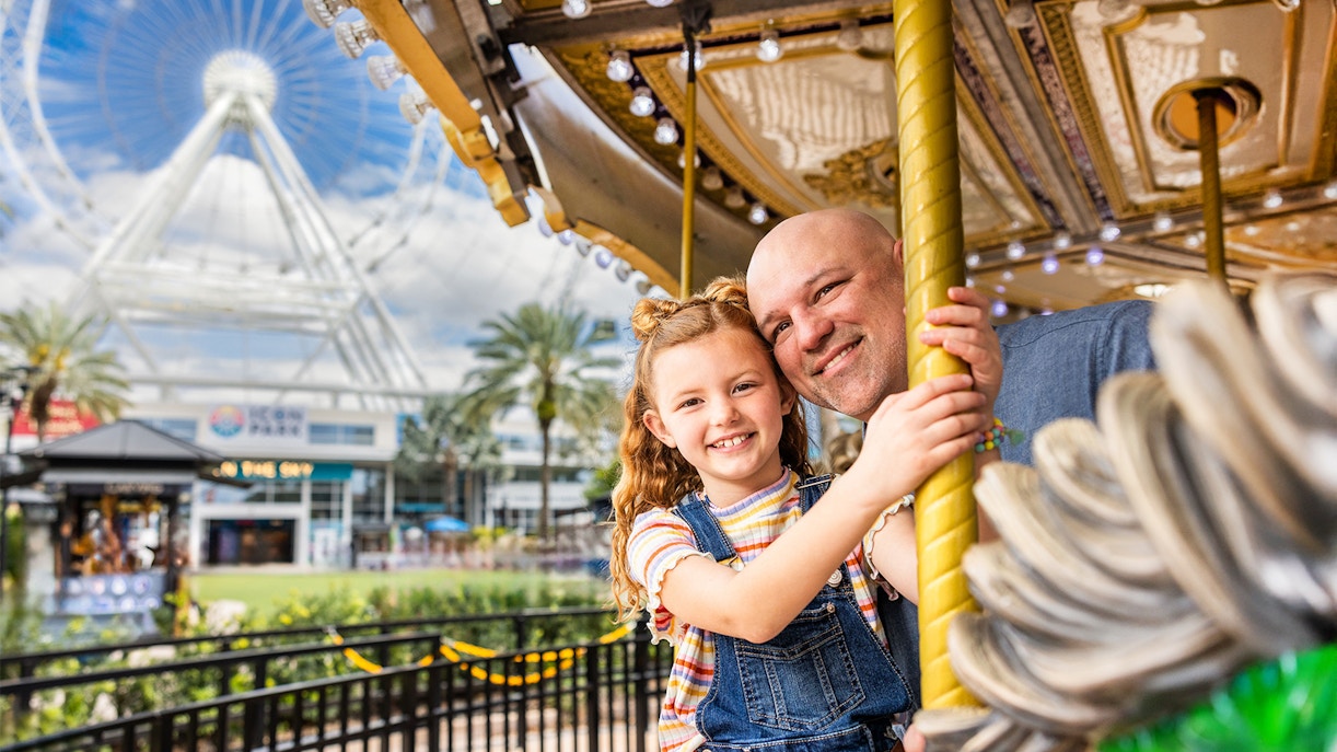 Father and daughter on a carousel at ICON Park, Orlando with The Wheel in the background.